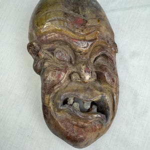 Puede incluir: Una máscara grotesca y desgastada con un acabado envejecido. La máscara presenta una boca ancha y abierta con dientes faltantes, cejas fruncidas y rasgos faciales exagerados. La paleta de colores incluye tonos de marrón, rojo y dorado.