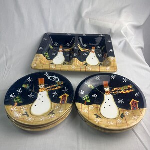 May include: A set of ceramic snowman-themed serving dishes. The set includes two round plates, a rectangular divided dish, and a snowman design with a black background, snowflakes, and a yellow and tan border.