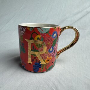 May include: A ceramic mug with a red background and a gold handle. The mug is decorated with colorful floral patterns and a large gold letter "R". The rim and handle are gold.