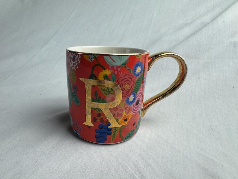May include: A ceramic mug with a red background and a gold handle. The mug is decorated with colorful floral patterns and a large gold letter "R". The rim and handle are gold.
