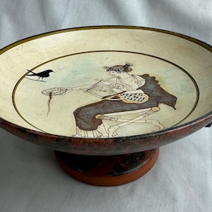 May include: A ceramic bowl with a painted classical scene. The bowl has a wide, shallow design with a brown rim and a circular base. The interior features a woman and a bird. The bowl is approximately 25 cm in diameter.