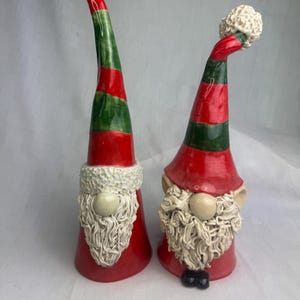 May include: Two ceramic gnome figurines. Each gnome has a red conical hat with green stripes, a white beard, and a beige nose. The hats are topped with a white, textured ball. One gnome has black shoes.
