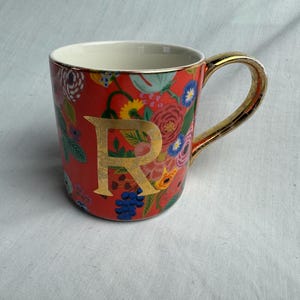 May include: A ceramic mug with a red background and a floral pattern in various colors. The mug features a gold handle and a gold letter "R". The interior of the mug is white.