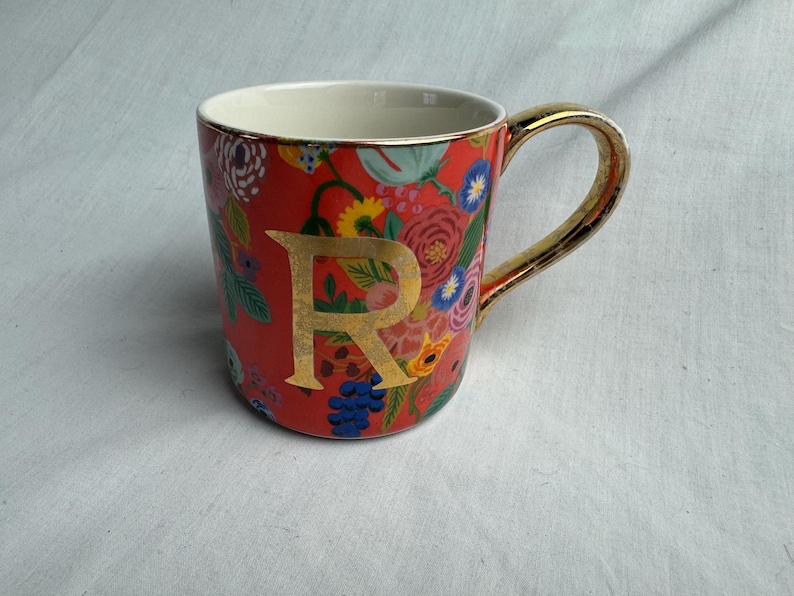 May include: A ceramic mug with a red background and a floral pattern in various colors. The mug features a gold handle and a gold letter "R". The interior of the mug is white.