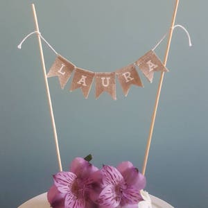 May include: A small cake topped with purple flowers and a banner that reads "LAURA". The banner is made of burlap and is strung between two wooden sticks. The cake sits on a white plate.