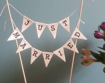 May include: A white cake topper with a banner that reads "JUST MARRIED" in black letters. The banner is strung between two wooden sticks. The cake topper is decorated with flowers.