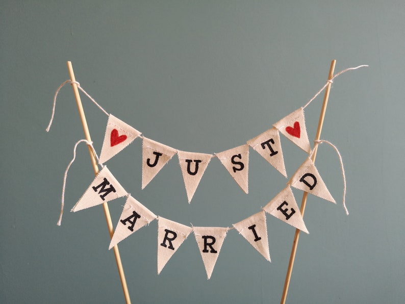 May include: A white fabric banner with the words "Just Married" in black lettering and red hearts. The banner is strung on a wooden dowel and is perfect for a wedding cake topper.