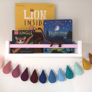 May include: A white shelf with three children's books on it. The books are "The Lion Inside", "Jungle", and "Night Time". There is a felt raindrop garland hanging below the shelf. The garland is made of felt raindrops in various colours, including pink, red, purple, brown, blue, and green.