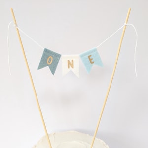 May include: A blue and white felt banner with gold letters spelling "ONE" on wooden sticks. The banner is perfect for a first birthday cake topper.