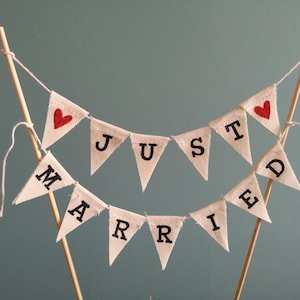 May include: A white fabric banner with red hearts and the words "JUST MARRIED" in black lettering. The banner is strung on a white string and is attached to two wooden sticks.