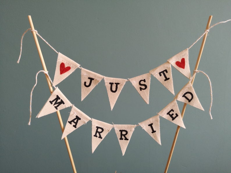 May include: A white fabric banner with red hearts and the words "JUST MARRIED" in black lettering. The banner is strung on a white string and is attached to two wooden sticks.