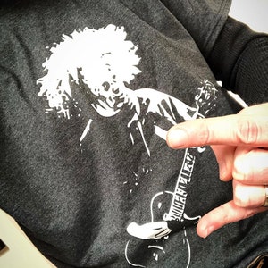 May include: A dark gray t-shirt with a white graphic print of a person with curly hair playing a guitar. The guitar has the text "Guitar Player" written on it.