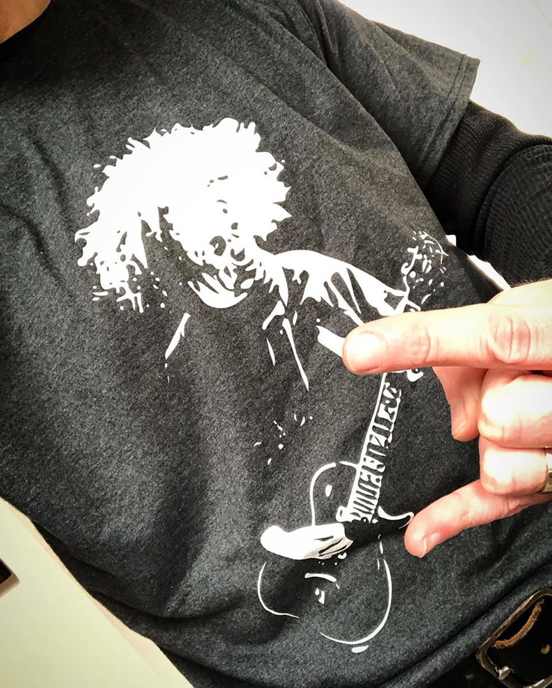 May include: A dark gray t-shirt with a white graphic print of a person with curly hair playing a guitar. The guitar has the text "Guitar Player" written on it.