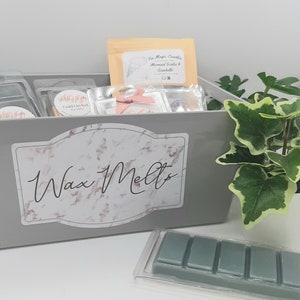 Puede incluir: Un contenedor gris con una etiqueta de m&aacute;rmol que dice "Wax Melts". El contenedor est&aacute; lleno de diferentes ceras para fundir, incluyendo un contenedor transparente con una etiqueta que dice "Gold Orchid Wax Melts". Tambi&eacute;n hay un paquete de ceras para fundir en un contenedor de pl&aacute;stico transparente con seis ceras para fundir individuales.