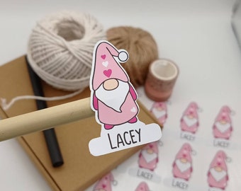 May include: Pink and white gnome sticker with a heart pattern and the name "Lacey" printed on a white banner. The sticker is on a brown cardboard box with a roll of twine and a roll of washi tape.