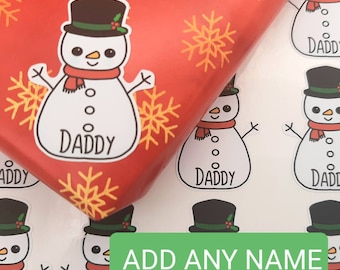 May include: Set of snowman gift tags with a red background and snowflakes. The snowman has a black top hat, a red scarf, and the text "Daddy" on the belly. The text "ADD ANY NAME" is visible in the image.