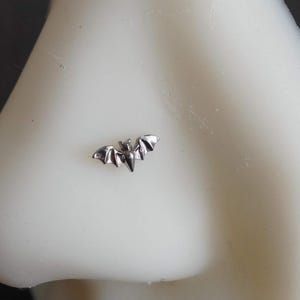 May include: A small, silver-colored bat-shaped nose stud jewelry piece. The bat is depicted with wings spread wide in a horizontal orientation, featuring detailed wing segments and a pointed ear design, positioned on a white nose model.