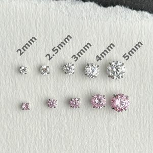 May include: A collection of titanium stud earrings in various sizes, ranging from 2mm to 5mm. The earrings are displayed on a white textured surface, with the word "titanium" printed above. Some earrings are clear, while others are pink.