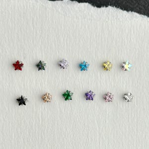 May include: A collection of small star-shaped stud earrings in various colors, including red, black, silver, blue, yellow, and iridescent. The earrings are displayed on a torn piece of white paper, showcasing their diverse hues and designs.