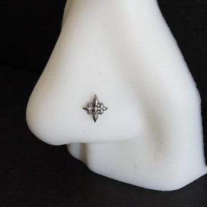 Könnte beinhalten: Ein silberfarbener Nasenstecker mit einem vierzackigen Stern- oder Lilien-Design, bestehend aus einem zentralen vertikalen Stab und symmetrischen seitlichen Akzenten, präsentiert auf einem weißen Nasenmodell zur Veranschaulichung.