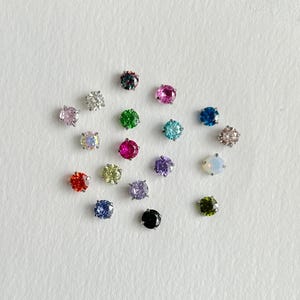 May include: A collection of small, round, faceted gemstones in various colors including red, blue, green, pink, purple, yellow, black, and white, each set in a metallic prong setting, arranged in a cluster on a textured white paper surface.