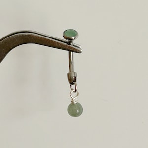 May include: A curved barbell body jewelry piece featuring a metallic silver-toned shaft with a small, smooth, pale green circular stone at the top and a matching pale green spherical bead dangling from a silver wire loop at the bottom end.