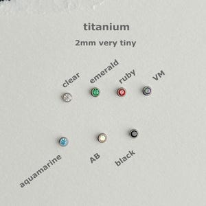 May include: A selection of 2mm titanium earrings in various colors, including clear, emerald, ruby, aquamarine, and black. The text on the image reads "titanium 2mm very tiny."