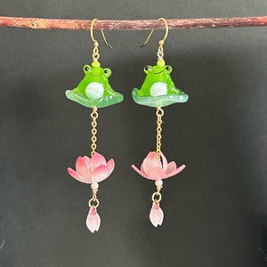 Peut inclure: Une paire de boucles d'oreilles pendantes avec des breloques de grenouilles vertes assises sur des nénuphars, avec des breloques de fleurs roses qui pendent en dessous.