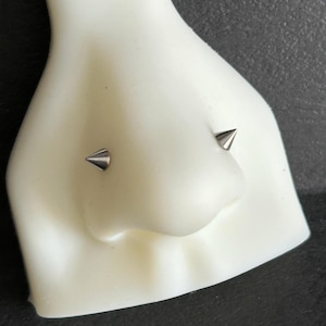 Pode incluir: Um expositor branco em forma de nariz com dois piercings pontiagudos prateados. Os piercings são simétricos e colocados nas narinas do expositor. O fundo é uma superfície texturizada cinza escuro.