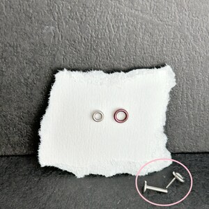 May include: A collection of small, silver-colored body jewelry pieces, including a flat-backed stud, a ring, and a barbell-style stud, are displayed on a white, torn piece of paper against a dark gray background.