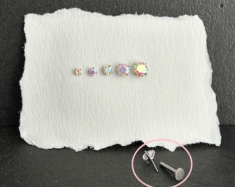 Pode incluir: Uma coleção de brincos de pressão redondos e iridescentes em vários tamanhos, exibidos em papel branco. Os brincos têm uma parte traseira plana e um pino para perfuração. Um único brinco é mostrado separadamente.