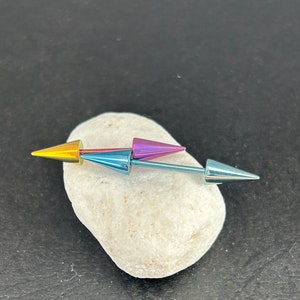 May include: A set of three colorful barbell piercings with pointed ends. The barbell is blue with a yellow tip on one end and a purple tip on the other end. The barbell is resting on a white rock.