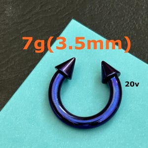 May include: A dark blue horseshoe piercing ring with pointed ends. The text "7g (3.5mm)" is above the ring, and "20v" is to the right. The jewellery is set against a light blue background.