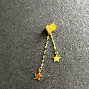 May include: A gold-colored earring with a rectangular top and two dangling chains, each ending in a small star. The earring is set against a dark gray background.