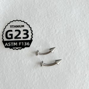 May include: Two silver-colored curved barbell body jewelry pieces with pointed cone ends. A black and white sticker is present with the text TITANIUM G23 ASTM F136, indicating the material composition of the metallic curved barbell jewelry.