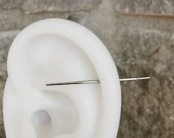 May include: A straight, silver-colored industrial barbell piercing jewelry featuring a long, thin metal bar that extends horizontally through a white silicone ear model, showcasing a smooth, polished finish and a simple, minimalist design.