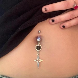 May include: A silver belly button ring with a dangling black heart and star charm. The top gem is a clear crystal, and the bottom charm is a four-pointed star. The ring is shown on a person's abdomen.