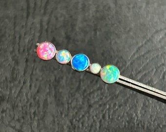 Könnte beinhalten: Eine Schmuckleiste aus Metall mit fünf kreisförmigen Cabochons in einer Reihe. Die Steine zeigen schillernde Farben, darunter Pink, Hellblau, leuchtendes Blau, Weiß und Grün. Das Stück wird von einer Metallpinzette gehalten und zeigt den Farbverlauf.