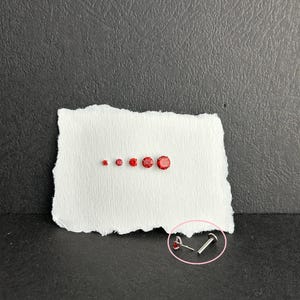 May include: A collection of small, round, red gemstones of varying sizes arranged on a white, torn piece of paper. A small, silver-colored stud earring with a red gemstone is also shown. The background is a dark gray color.