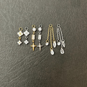 May include: A collection of six metal jewelry charms in gold and silver tones, featuring clear crystal accents. The set includes two flower-shaped charms, two star-shaped charms with rectangular crystals, and two multi-chain charms with teardrop crystals.