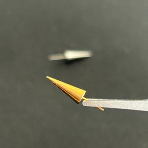 May include: A small, gold-coloured, conical object is held by tweezers against a dark background. The object has a pointed tip and a cylindrical base. Another, similar object is blurred in the background.
