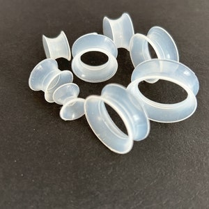 May include: A collection of translucent, light blue silicone ear gauges in various sizes. The gauges have a tunnel design, with a rounded outer edge and a flared inner edge. They are arranged on a dark surface.