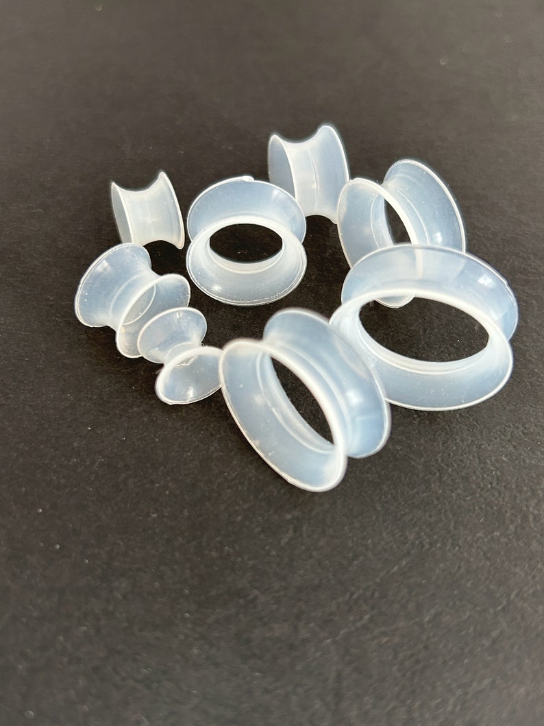 May include: A collection of translucent, light blue silicone ear gauges in various sizes. The gauges have a tunnel design, with a rounded outer edge and a flared inner edge. They are arranged on a dark surface.