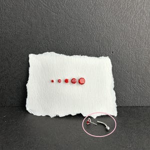 May include: A selection of small, round, red gemstones of different sizes on white paper. A curved, silver-coloured body jewellery piece with a small red gemstone is also present. The background is a dark grey surface.