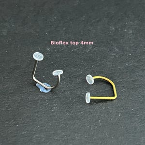 Può includere: Due piercing al naso con estremità trasparenti. Uno è color argento, l'altro dorato. Il testo "Bioflex top 4mm" è visibile. I piercing sono su una superficie scura.