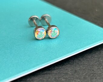 Pode incluir: Dois brincos de botão prateados com pedras preciosas redondas e iridescentes. Os brincos são exibidos em uma superfície azul-turquesa. Cada brinco tem uma parte traseira plana e um pino para perfurar.