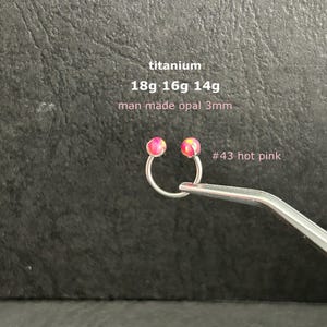 May include: A circular titanium body jewelry piece with two 3mm hot pink man-made opal beads. Text on the image reads: titanium, 18g 16g 14g, man made opal 3mm, #43 hot pink. The jewelry is held by metal tweezers against a dark textured background.