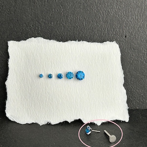 May include: An assortment of blue gemstone studs in different sizes, presented on a torn piece of white paper. The studs are set against a dark backdrop. One stud is shown with its post and backing.