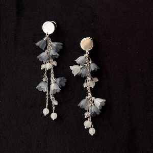 May include: Dangle earrings with silver-tone metal discs and chains. Each earring features blue-grey flower charms and small white beads. The earrings have a delicate, floral design. The background is black.