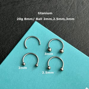 May include: Four silver titanium horseshoe-shaped body jewellery pieces with ball ends. The text on the image reads "titanium 20g 8mm/ Ball 2mm,2.5mm,3mm". The jewellery is displayed on a teal blue surface.